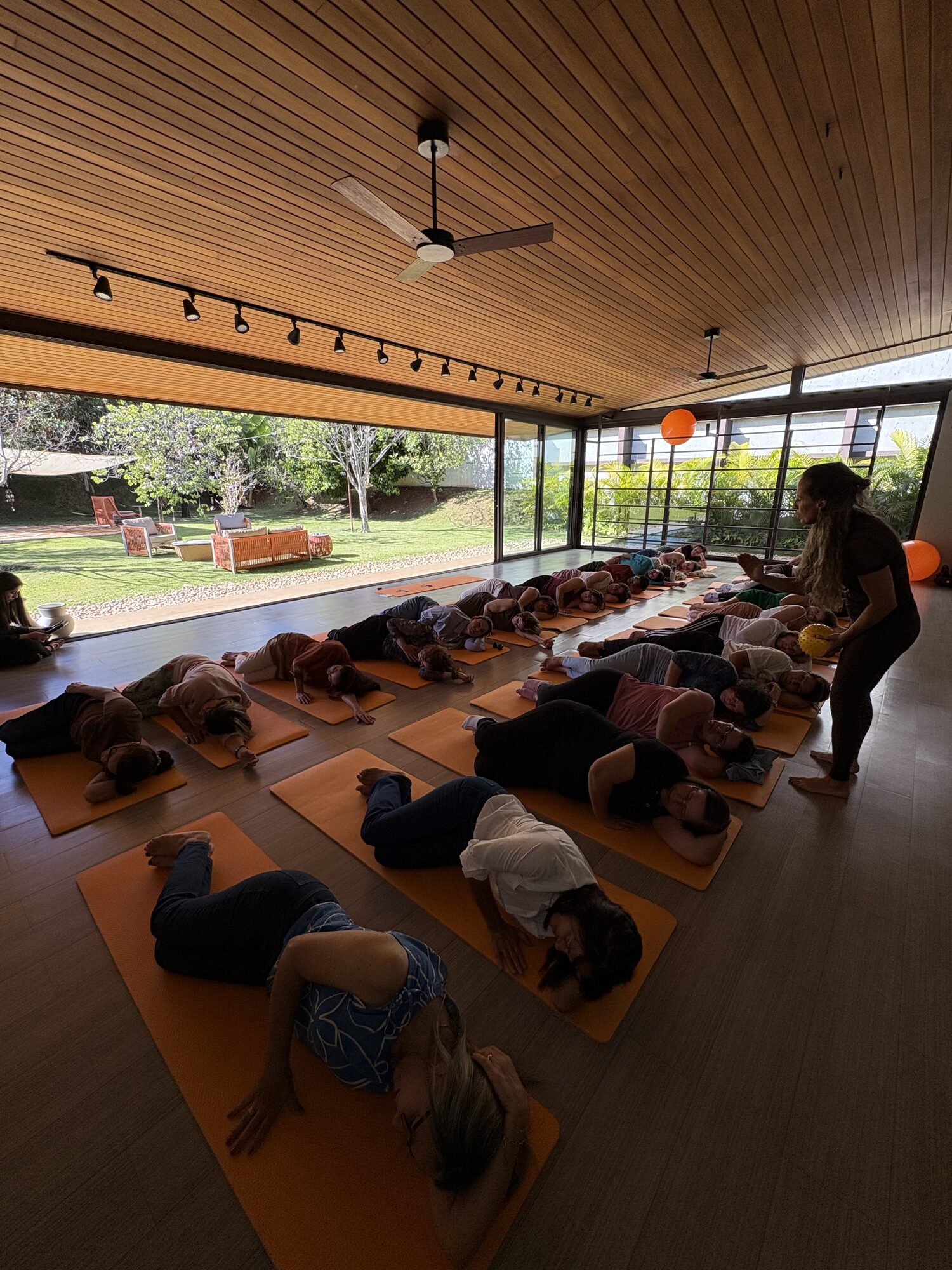 Dezenas de mulheres em savasana sobre tapetes laranjas no estúdio de yoga da Casa Aromamundo, com vista para o jardim